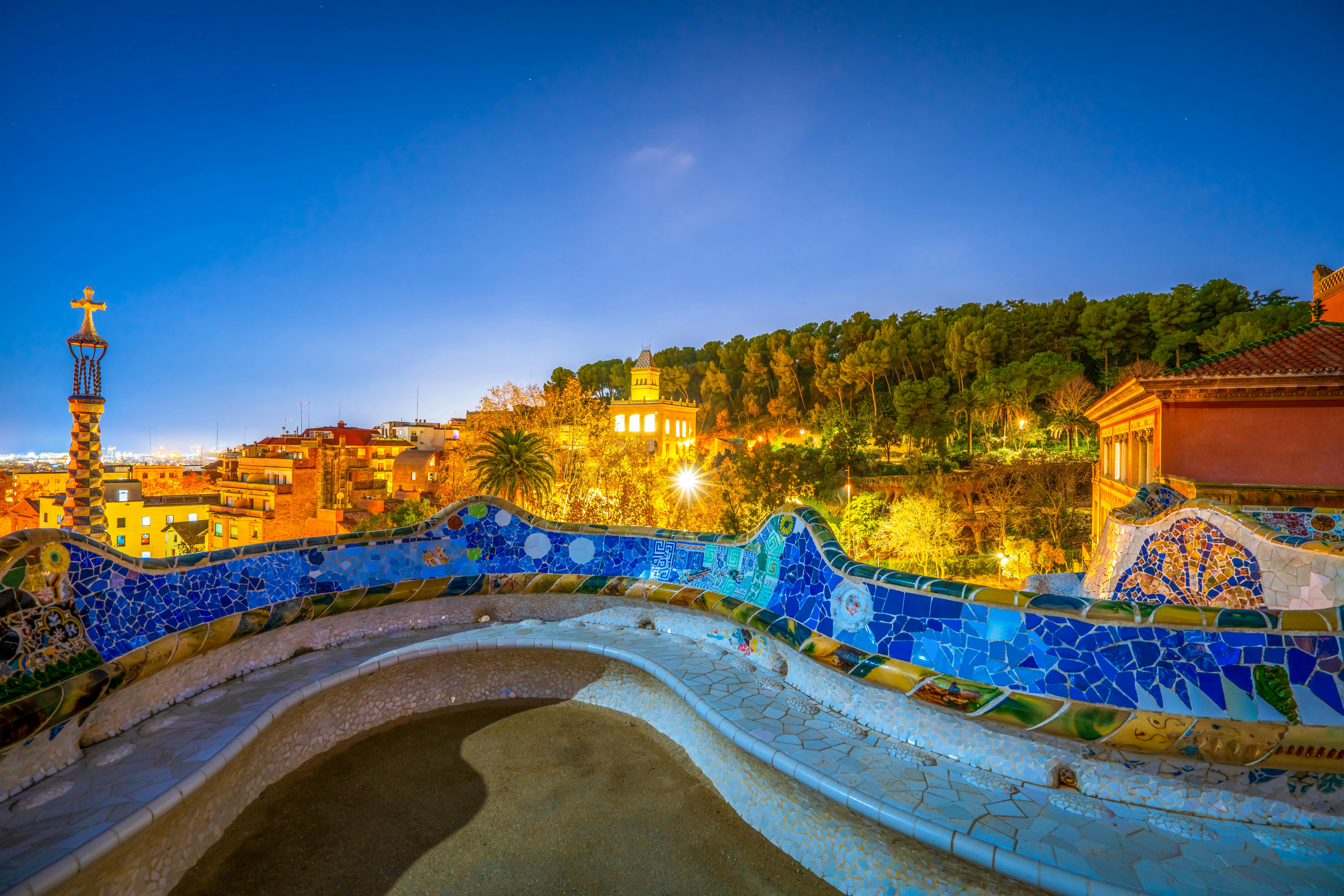 Banc en mosaïque incurvée surplombant un parc et un bâtiment illuminé sur fond d'arbres et de ciel crépusculaire.