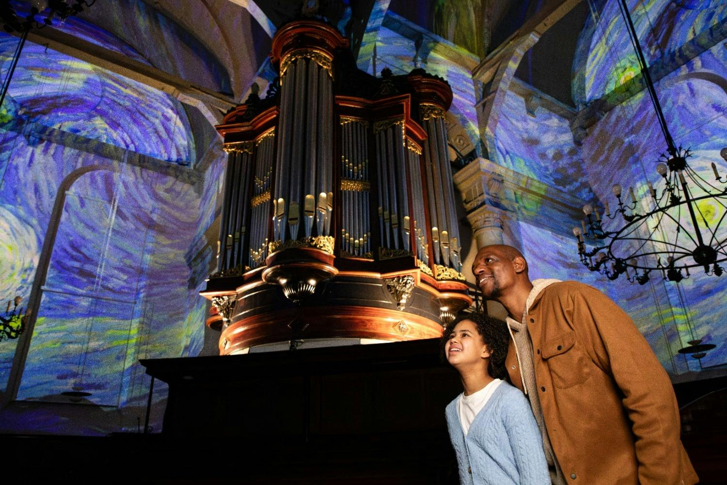 Δύο άνθρωποι θαυμάζουν ένα μεγάλο εκκλησιαστικό όργανο σε ένα φωτισμένο εσωτερικό με πολύχρωμες καλλιτεχνικές προβολές στους τοίχους και την οροφή.
