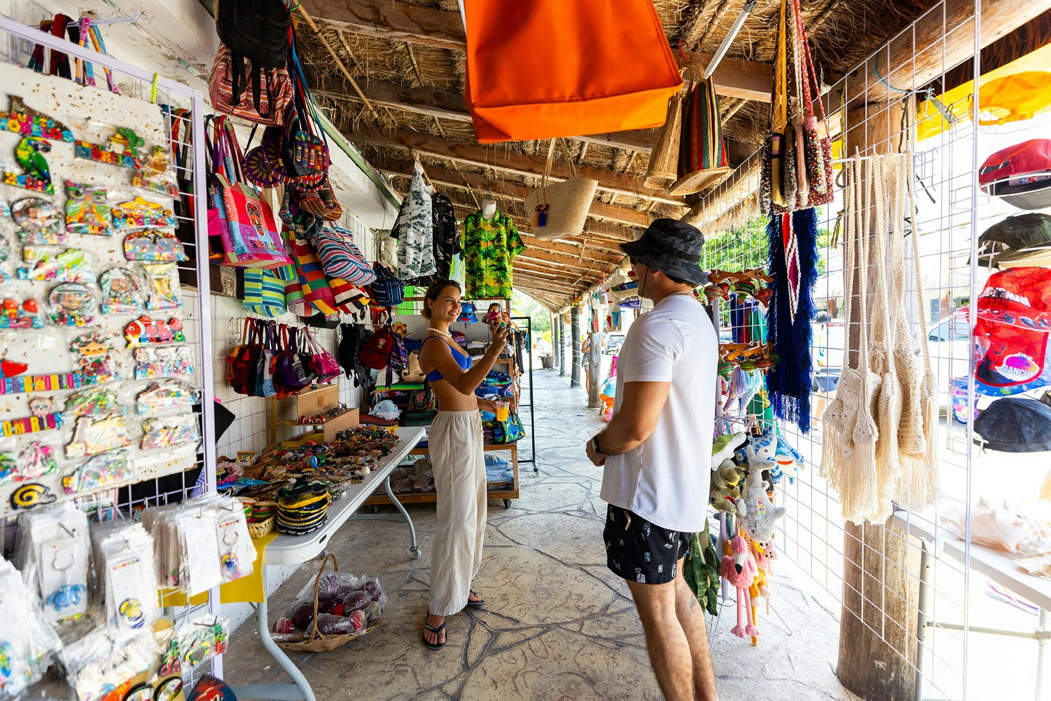 Ein Mann und eine Frau stöbern auf einem farbenfrohen Markt im Freien, auf dem verschiedene Taschen, Kleidung und Accessoires zum Verkauf angeboten werden.