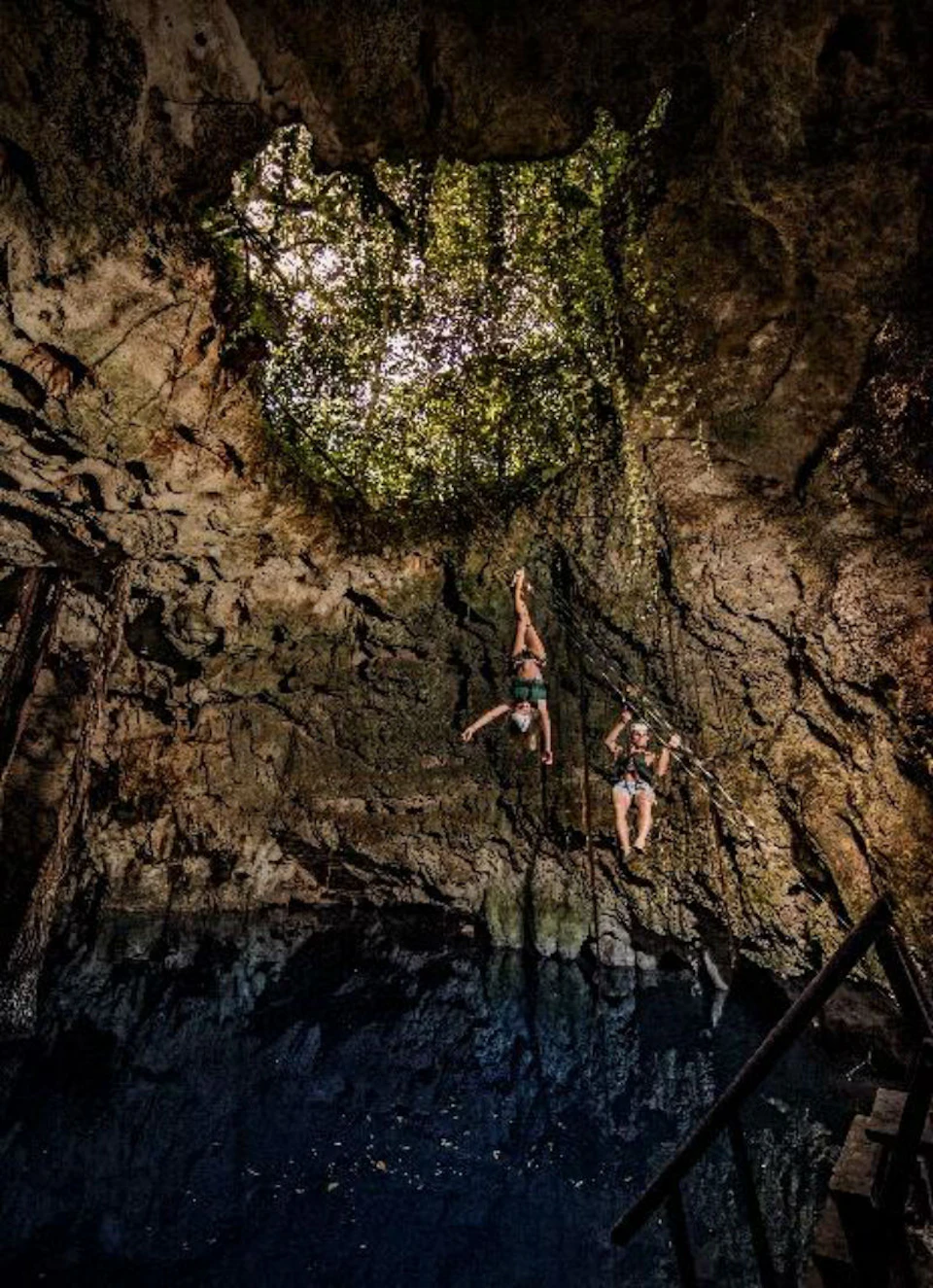Two people hang on ropes inside a cave with an opening at the top, illuminated by sunlight filtering through foliage.