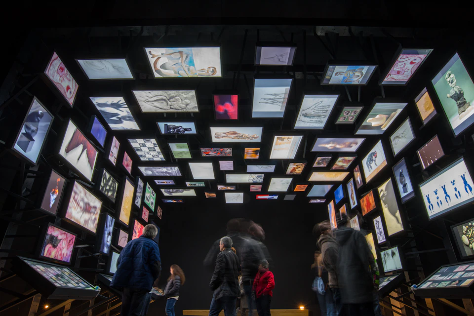 People observe a dark room with illuminated art panels displayed on the walls and ceiling, creating a vibrant visual experience.