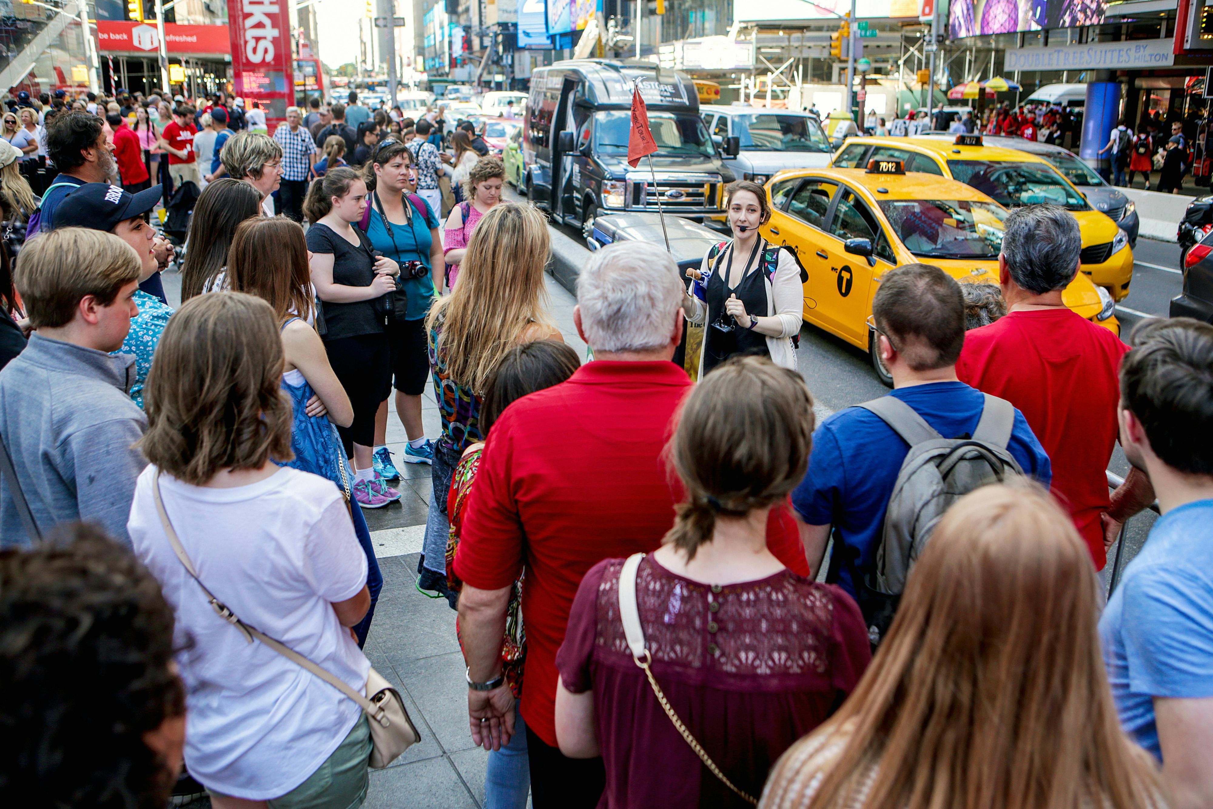 A tour guide speaks to a group of people on a busy city street with yellow cabs and cars in the background.