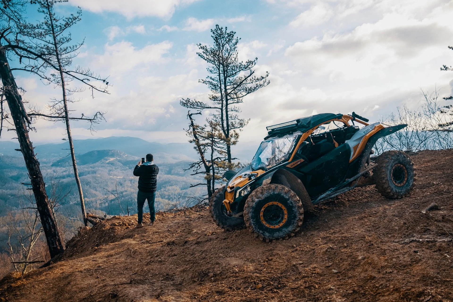 A person taking a photo near an off-road vehicle on a dirt hill with a scenic mountain view and trees in the background.