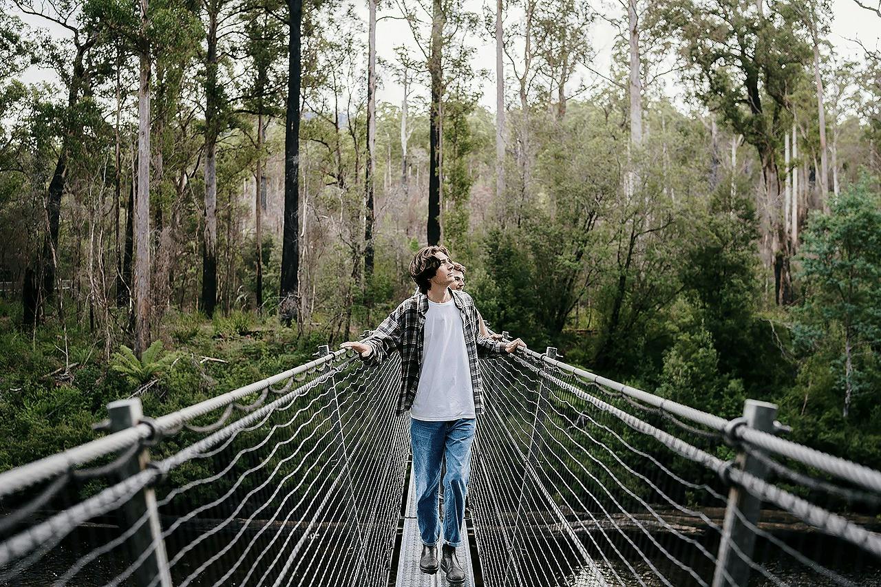 A person stands on a suspension bridge in a forested area, holding onto the railing, with trees visible in the background.