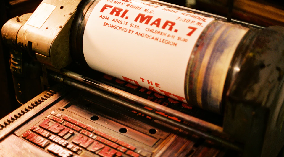 Close-up of a printing press rolling a poster that reads 'FRI, MAR, 7' and other event details.