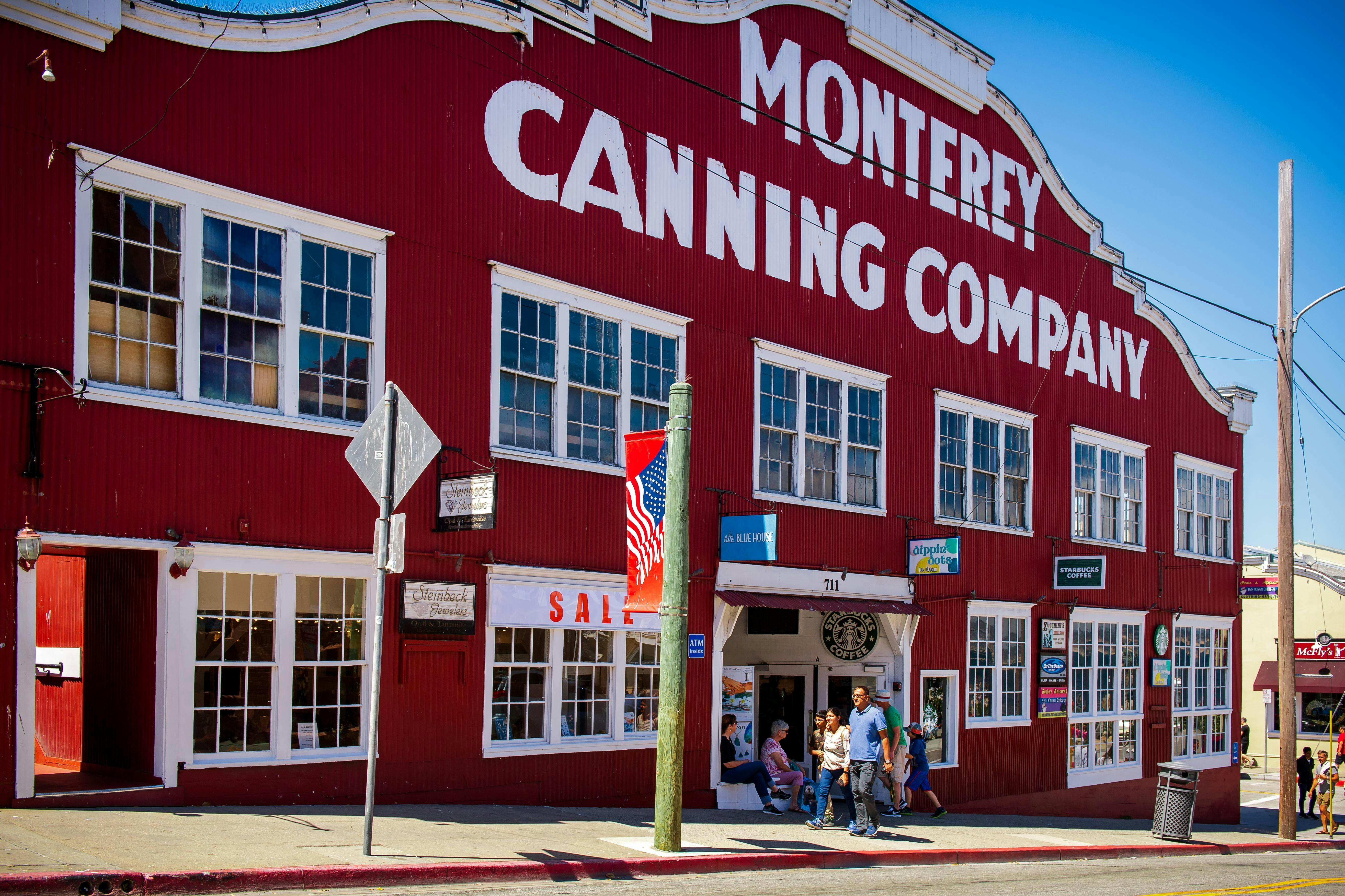 Ένα κόκκινο κτίριο με την ένδειξη "Monterey Canning Company" με ανθρώπους να περπατούν δίπλα στην είσοδο και μια αμερικανική σημαία να εμφανίζεται έξω.