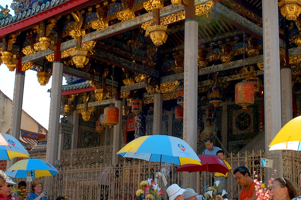 People riding colorful trishaws with umbrellas in front of an ornate, traditional temple building under a clear sky.