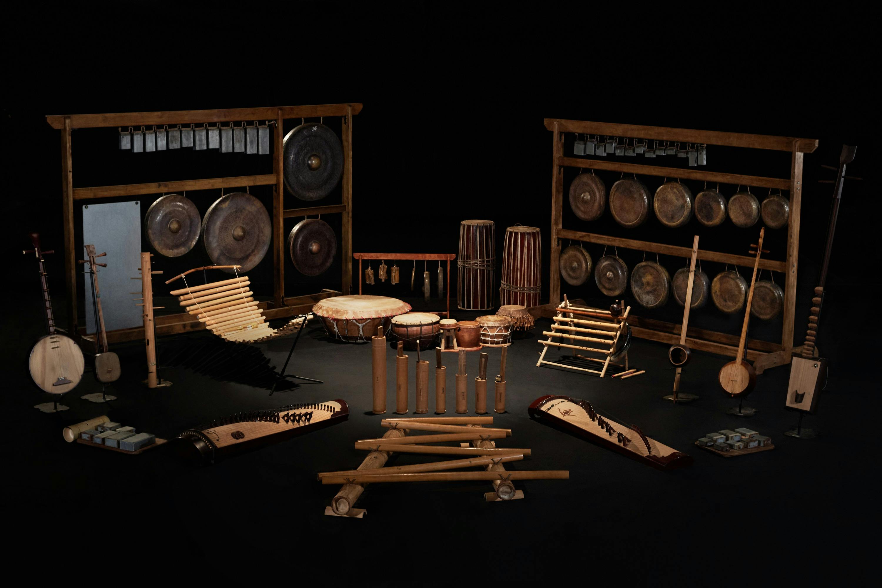 A collection of traditional musical instruments, including gongs, drums, xylophones, and string instruments, on a dark background.