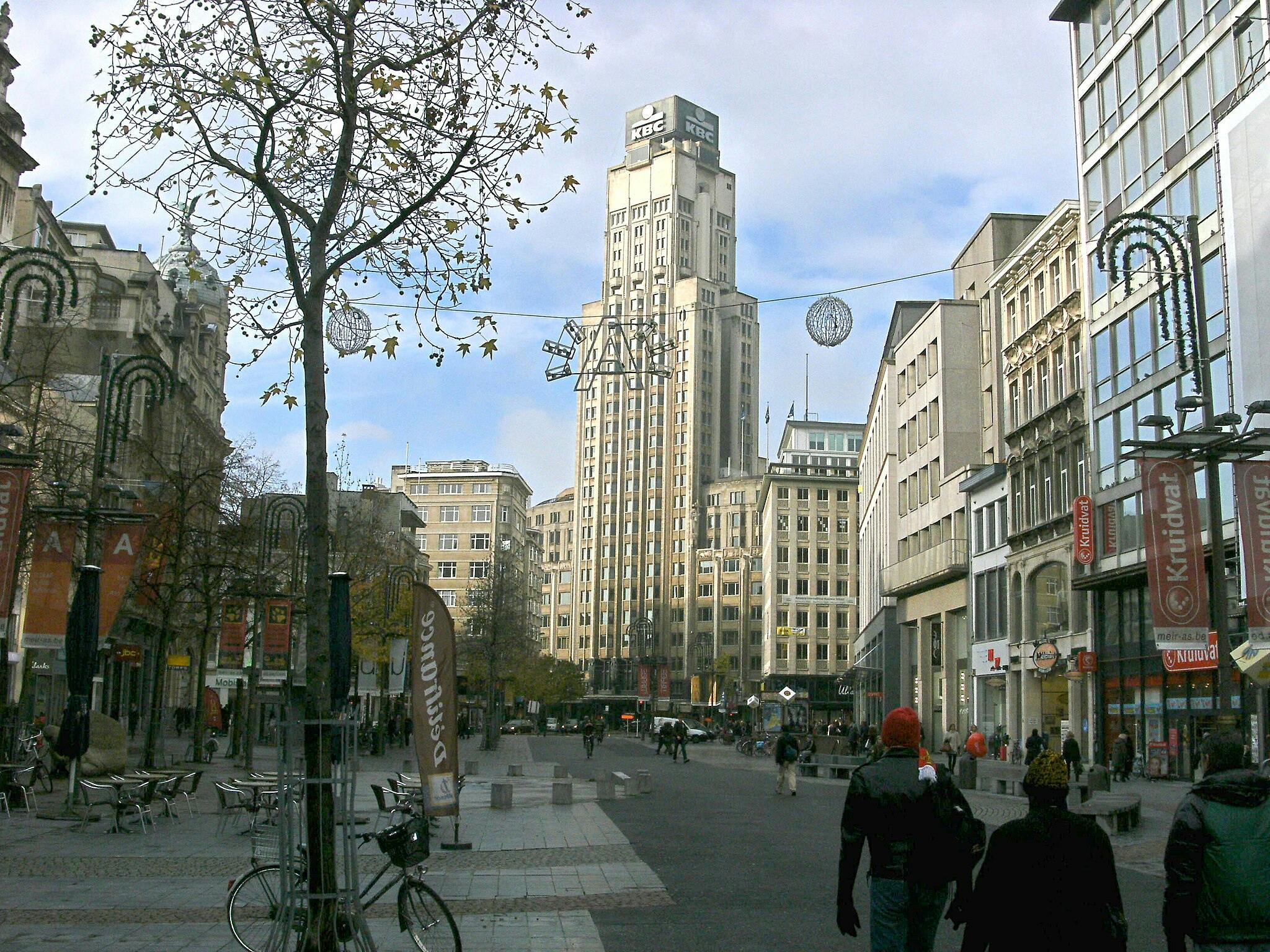 People walk through a city street lined with trees and buildings, featuring a tall tower in the background and festive decorations.