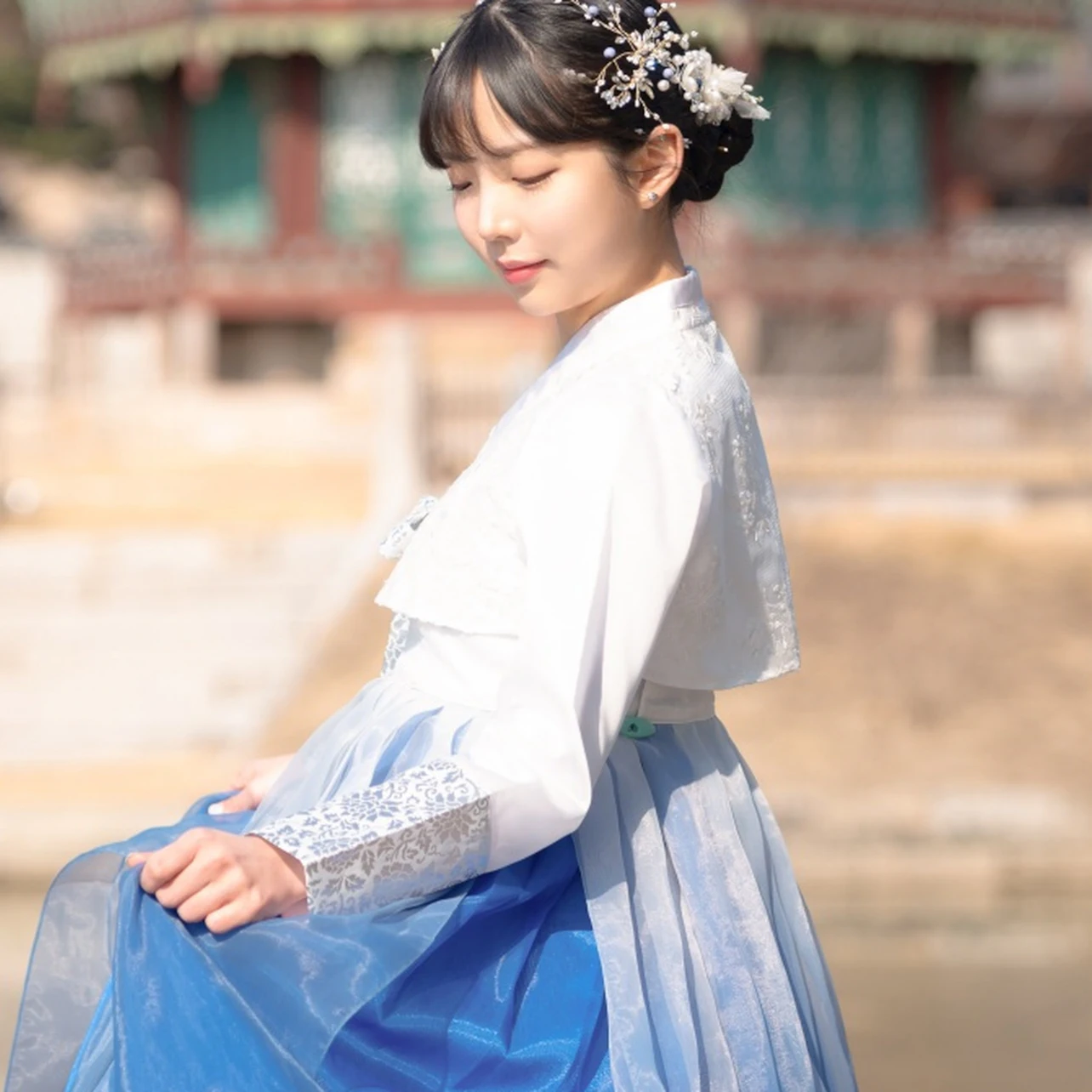 A woman in traditional attire poses gracefully outdoors, with a historical building in the background.