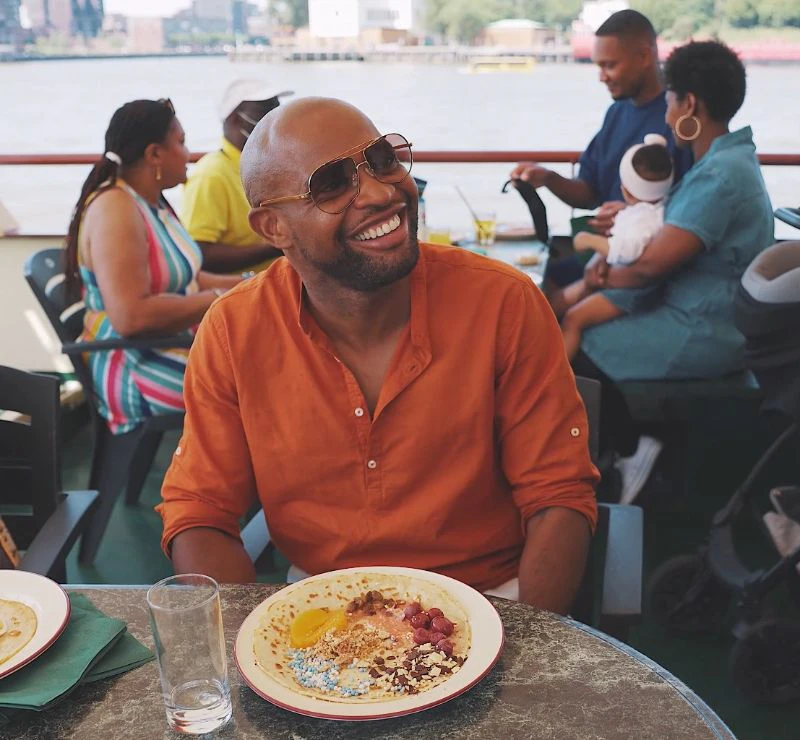 A smiling person in an orange shirt sits at a table with a partially eaten meal. Others are seated around, holding a baby.