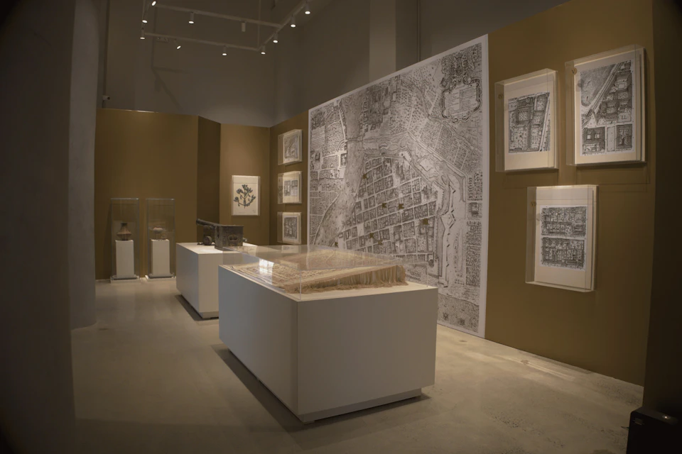 An exhibition space displaying maps and architectural models on walls and pedestals under soft lighting.