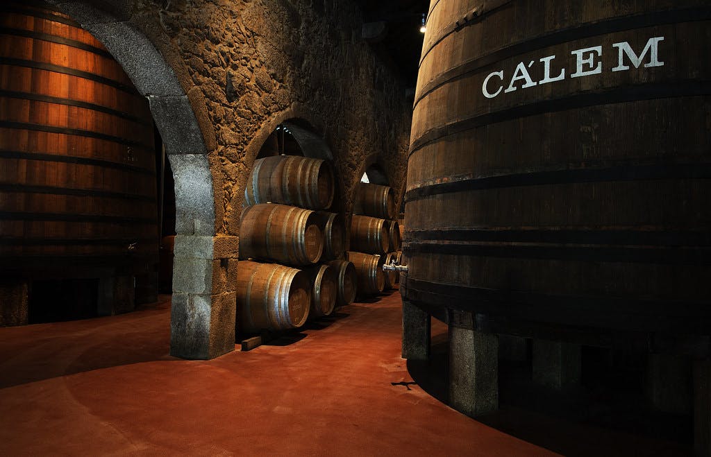 Interior of a wine cellar with large wooden barrels, stacked smaller barrels, and stone archways under dim lighting.