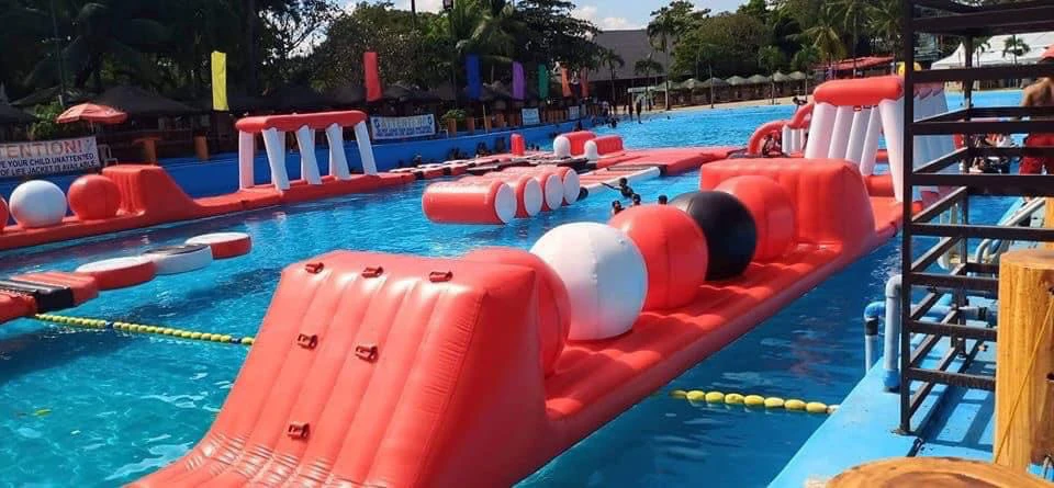 An inflatable obstacle course floating on a swimming pool, with red, white, and black sections and people in the background.