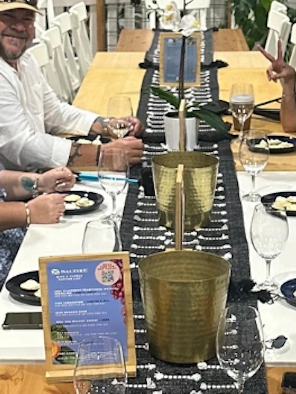 A group of people seated at a long table in a winery setting, with tanks in the background and food on the table.