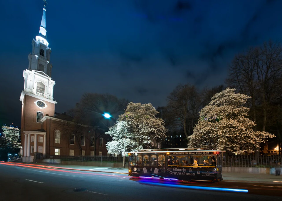 A tour bus labeled "Ghosts & Gravestones" moves past a church with a lit steeple and flowering trees at night.