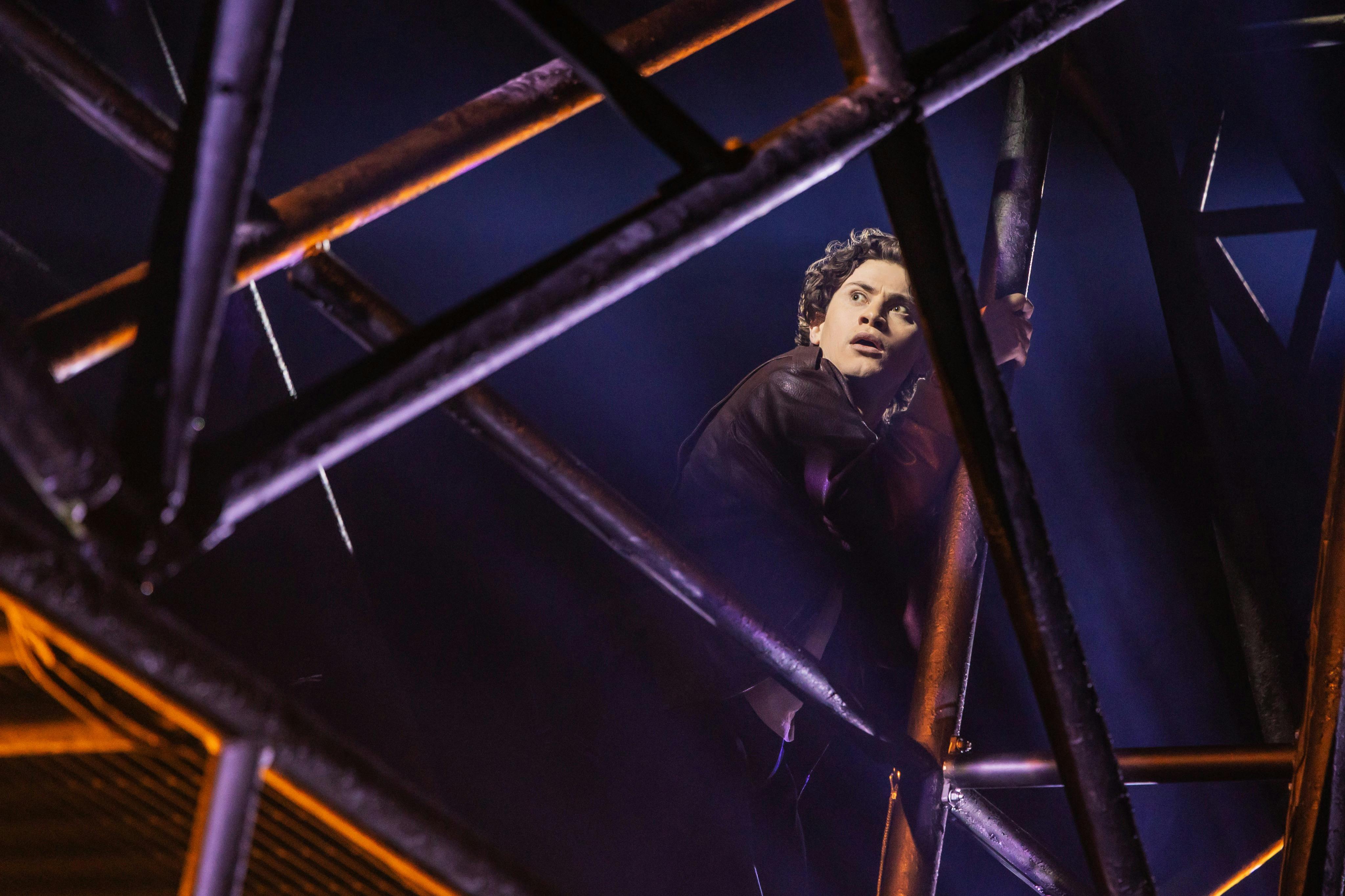 A person with curly hair clings to a metal structure, illuminated by dramatic lighting, with a dark and smoky background.