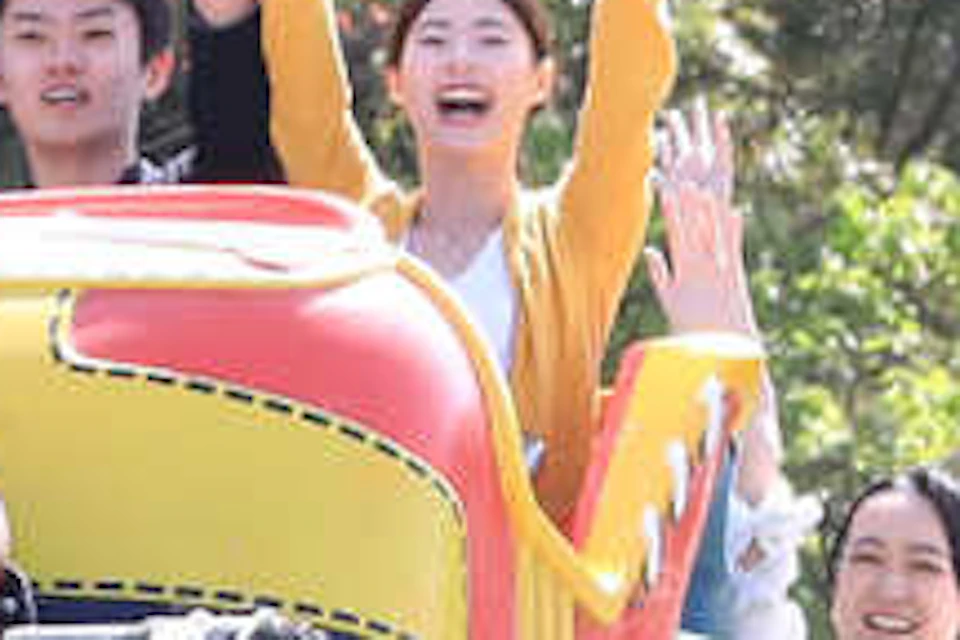 A colorful roller coaster with passengers raising their hands in excitement, surrounded by green trees on a sunny day.