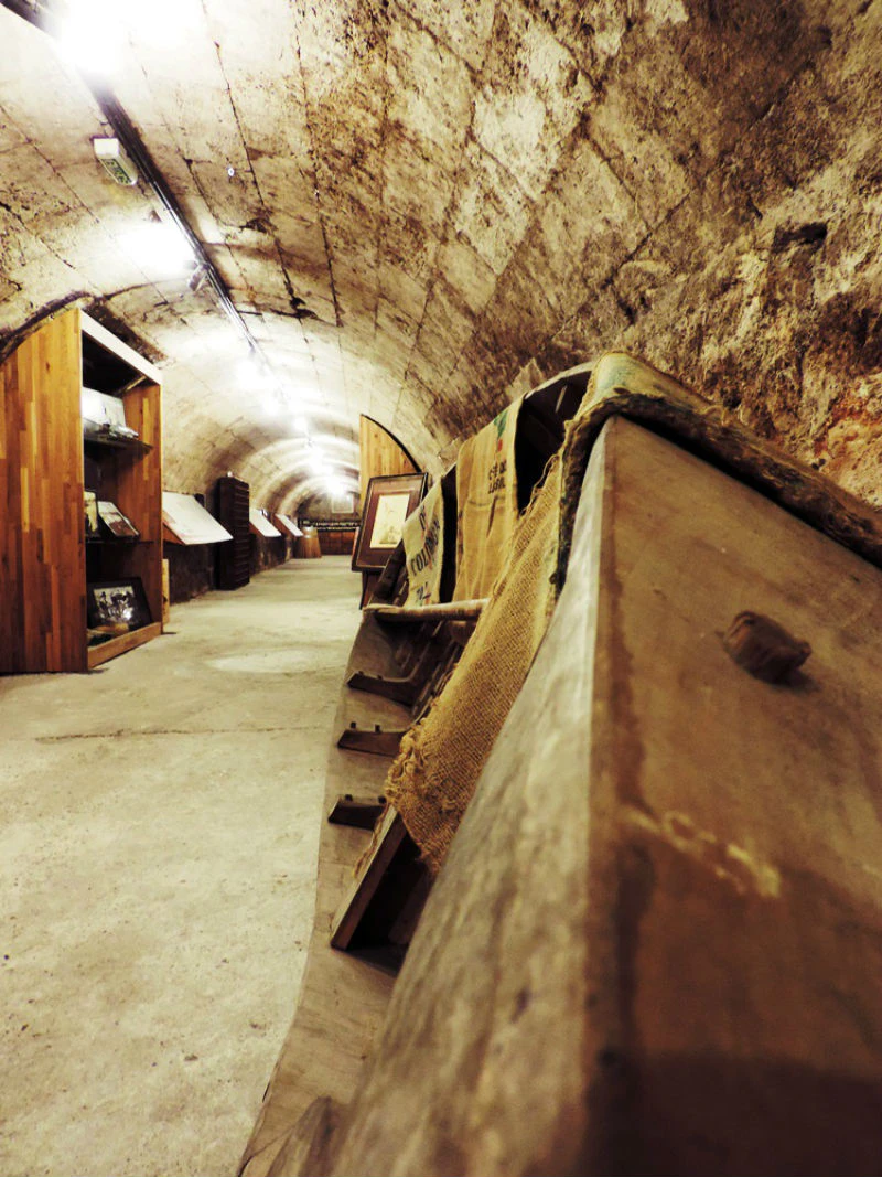 An underground tunnel with wooden displays on both sides, old wooden barrels, and burlap sacks lining the walls.