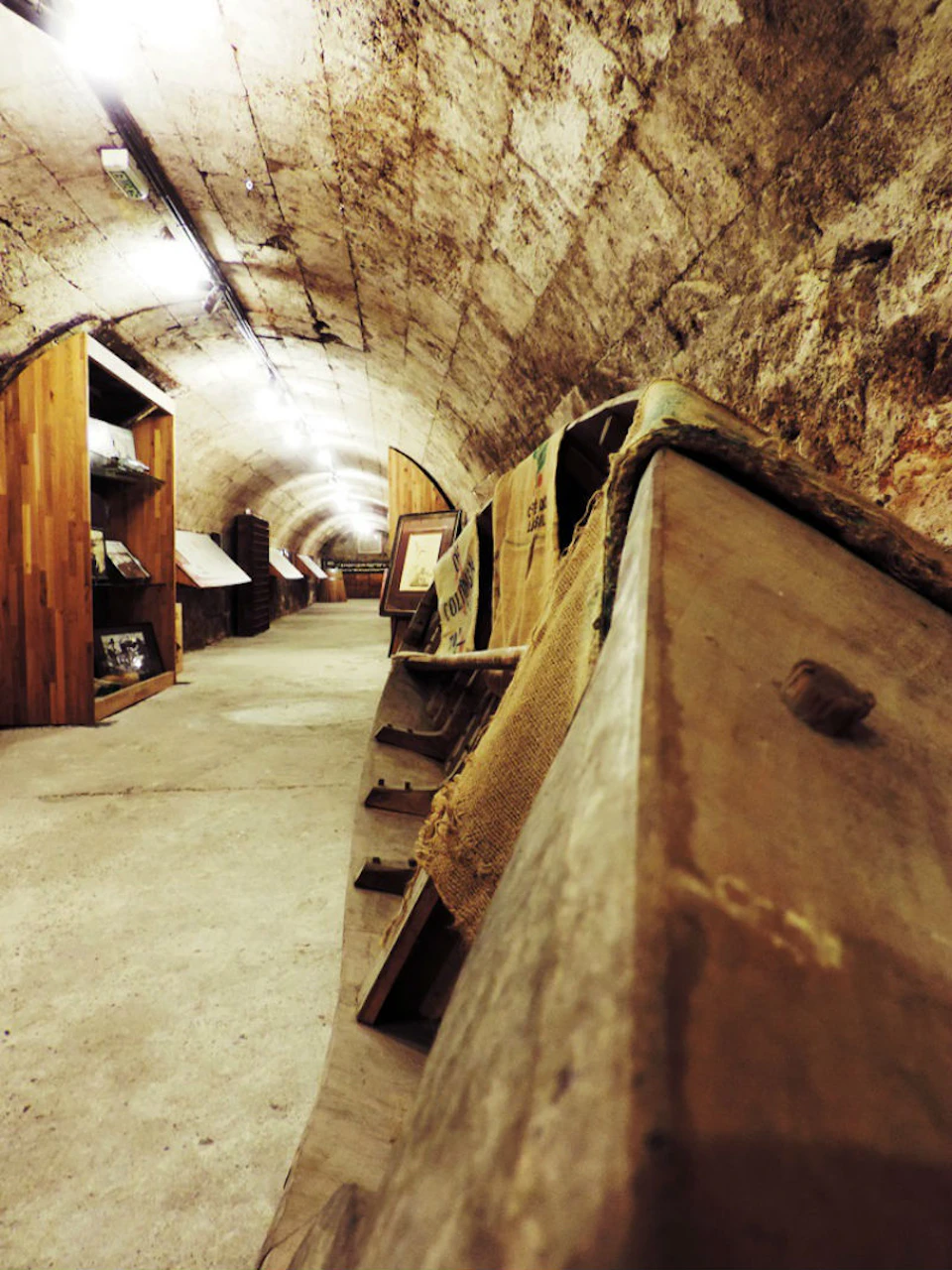 An underground tunnel with wooden displays on both sides, old wooden barrels, and burlap sacks lining the walls.