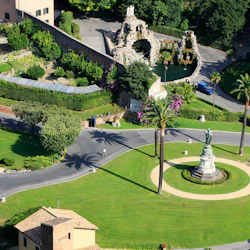 Flygfoto över en anlagd trädgård med en central staty, böjda gångar, palmer, buskar och ett litet fontänområde.