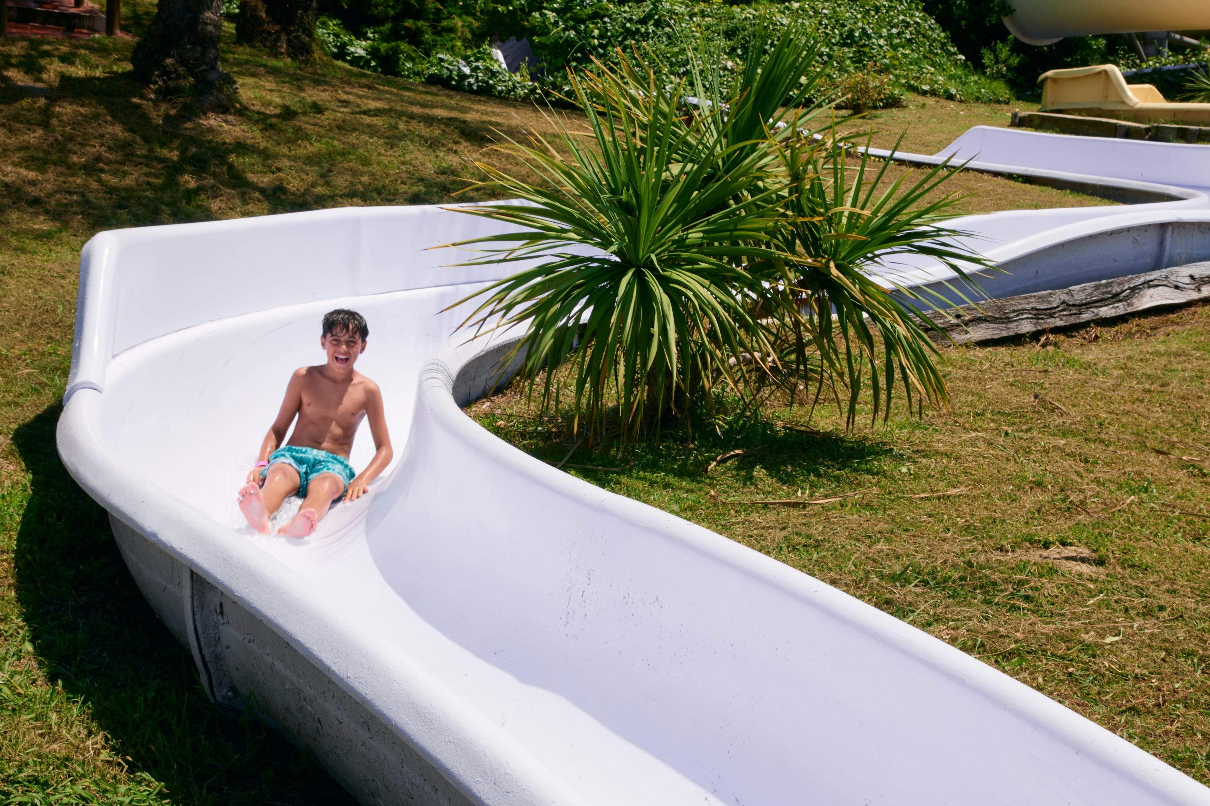 A boy sliding down a white water slide outdoors surrounded by greenery on a sunny day.
