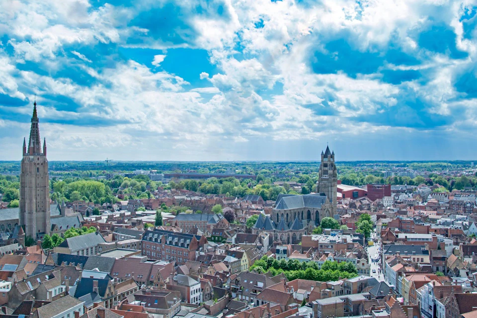 Aerial view of a European city with red-roofed buildings, a large church, and surrounding greenery under a partly cloudy sky.