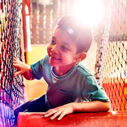 Leende barn i en grön skjorta leker i en nätad lekplats med starkt solljus i bakgrunden.