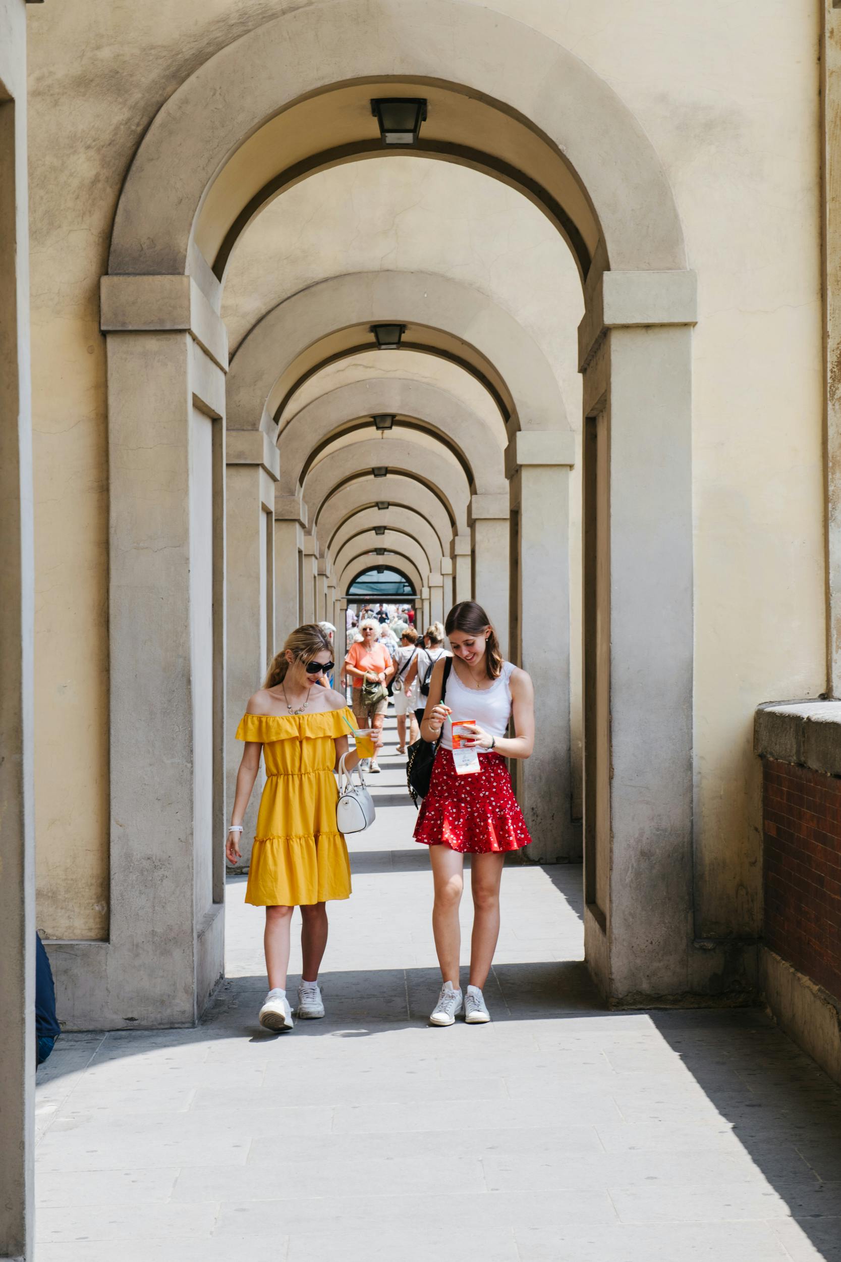 Δύο γυναίκες περπατούν κάτω από πέτρινες καμάρες. Η μία φορά κίτρινο φόρεμα και η άλλη κόκκινη φούστα. Άνθρωποι και φως ορατό στο παρασκήνιο.