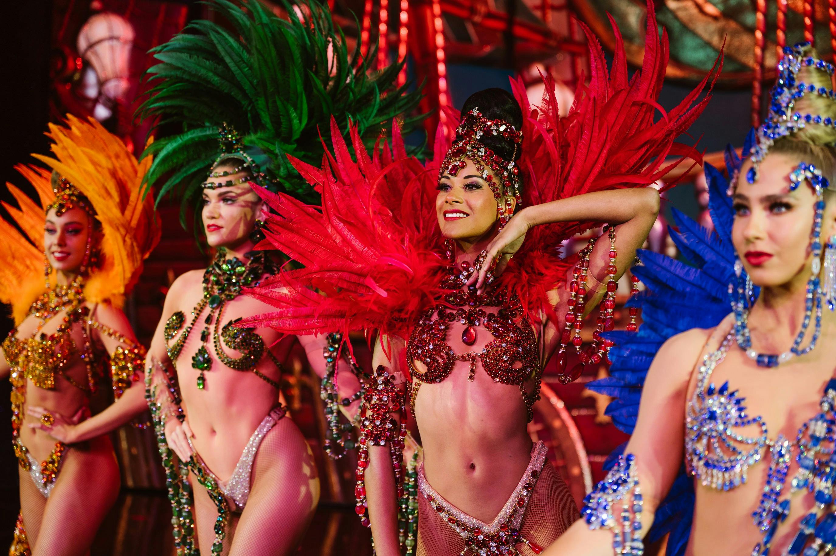 Performers in colorful feathered costumes and ornate accessories dancing on a brightly lit stage.