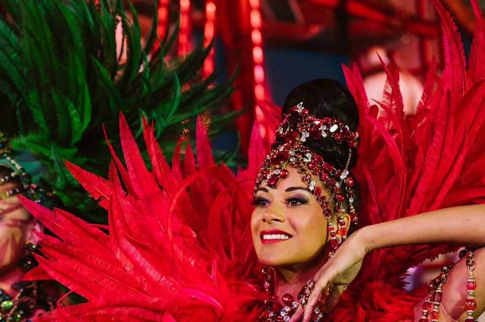Performers in colorful feathered costumes and ornate accessories dancing on a brightly lit stage.