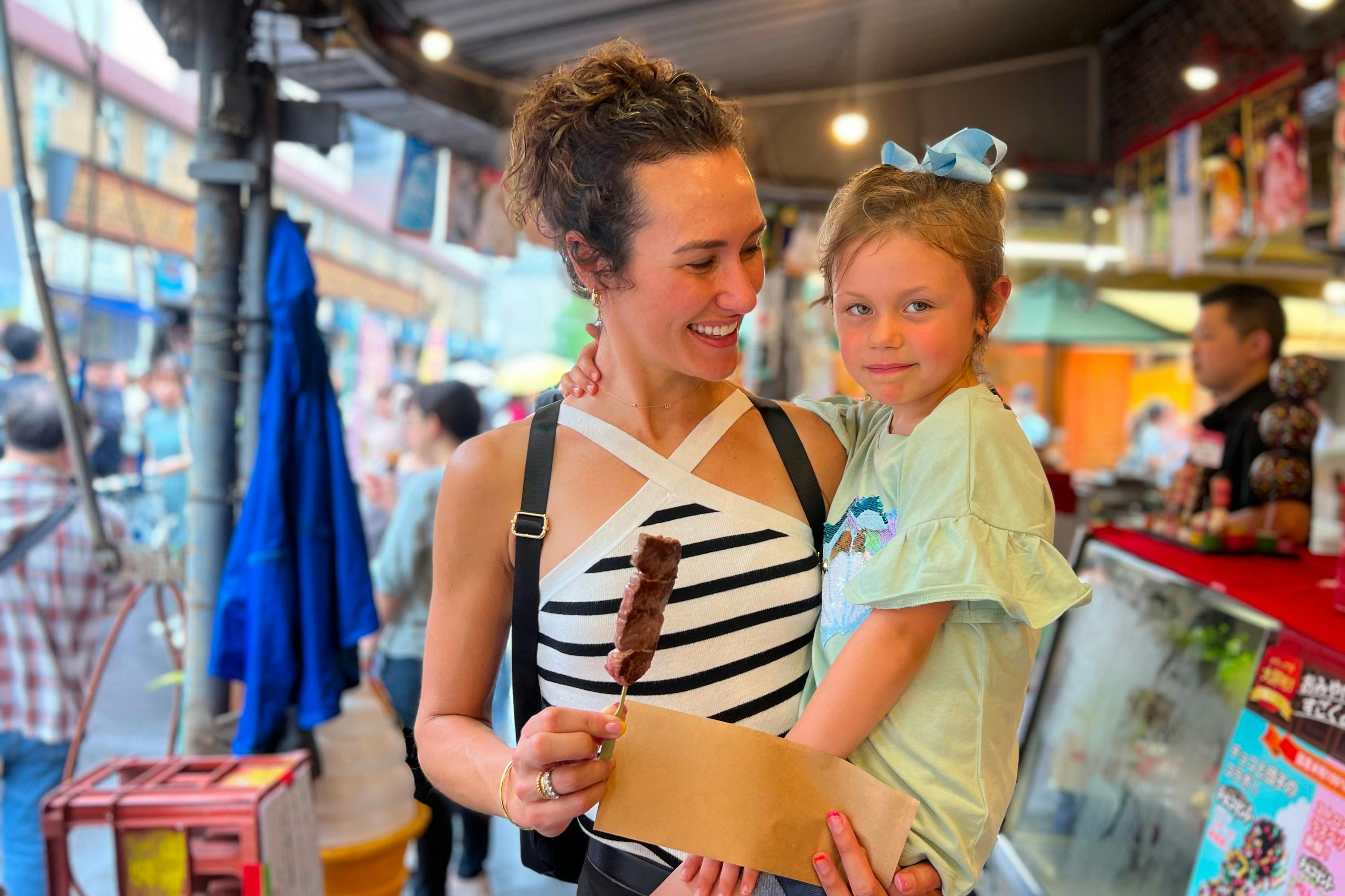 A woman smiling at a young girl while holding a chocolate-covered treat on a stick, in a lively marketplace setting.