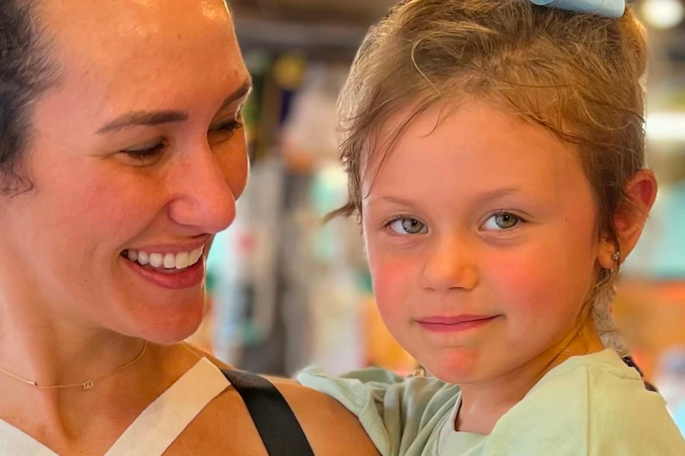 A woman smiling at a young girl while holding a chocolate-covered treat on a stick, in a lively marketplace setting.