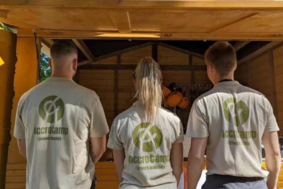 Three people with "AccroCamp" shirts stand facing an open wooden storage shed with climbing gear and helmets.