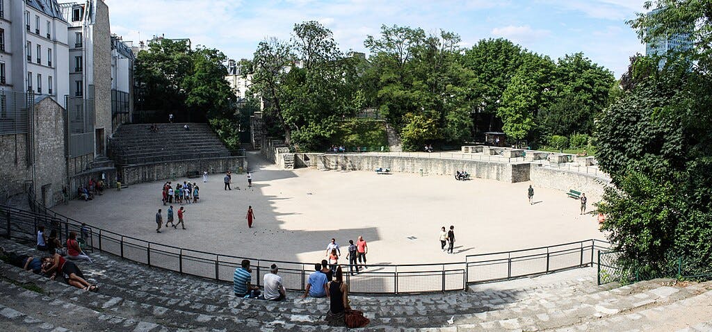 People sitting and walking in a historic stone amphitheater surrounded by trees and buildings on a sunny day.