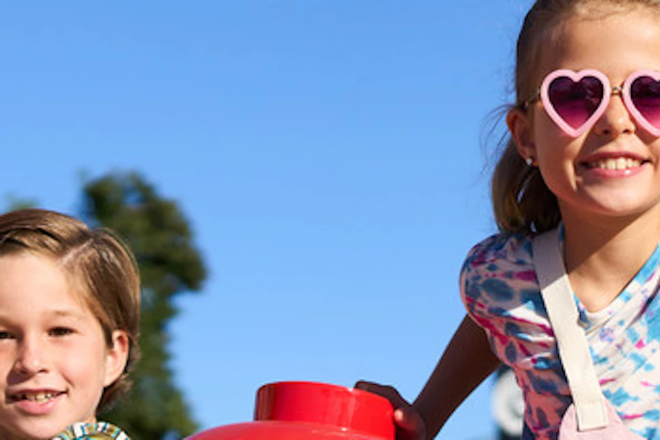 Two children smiling and playing on colorful playground equipment, with oversized red, green, and yellow fruit sculptures around them.