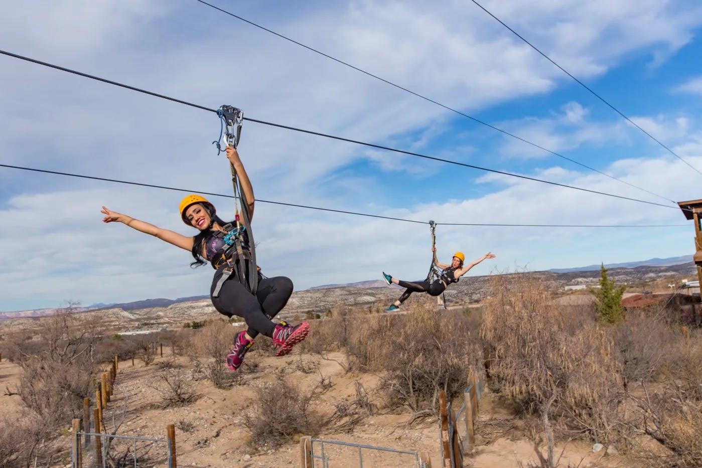 Два человека застегиваются на молнию над суровым пустынным ландшафтом, каждый из которых одет в шлемы и ремни безопасности под частично облачным небом.