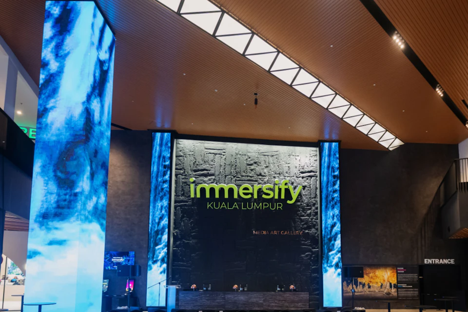 A media art gallery entrance with the text 'Immersify Kuala Lumpur' on the wall, flanked by large blue-lit columns.