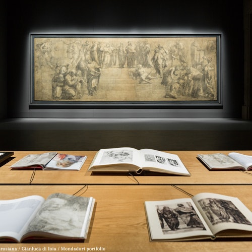 Open books with sketches on a wooden table, set in front of a large framed grayscale artwork displayed on a dark wall.