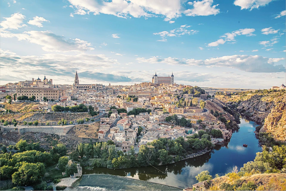 A scenic view of a historic city with a river, numerous buildings, and a large cathedral under a partly cloudy sky.
