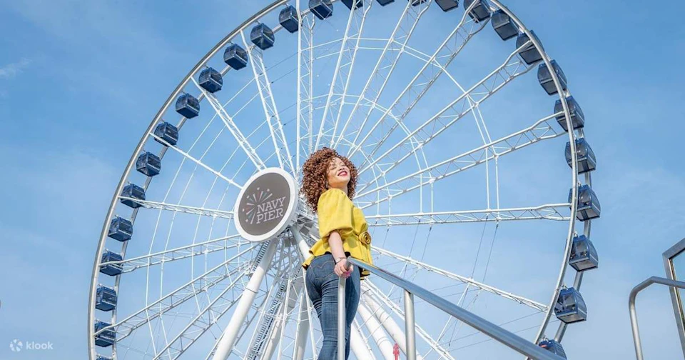 A smiling woman stands in front of the Navy Pier Ferris wheel, wearing a yellow jacket and jeans on a sunny day.
