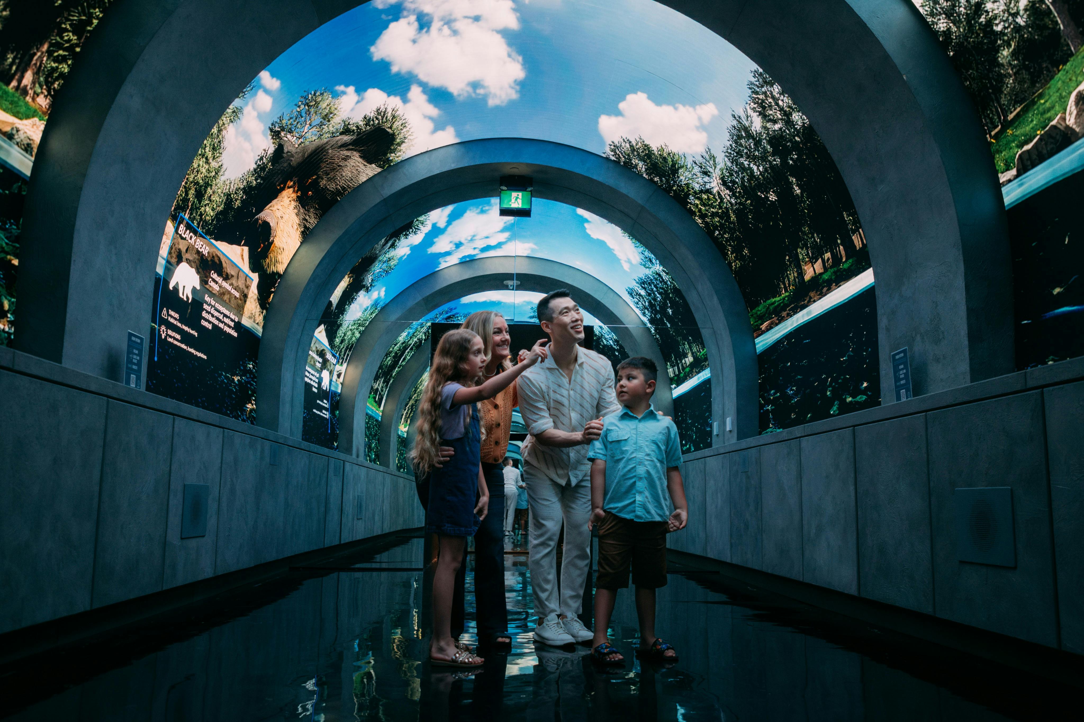 A family of four stands under an arched digital display showing trees and sky, smiling and pointing.