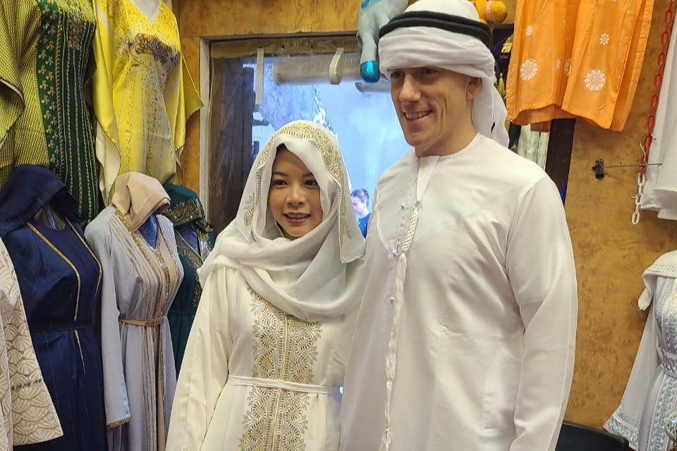 Ένας άνδρας και μια γυναίκα ντυμένοι με παραδοσιακά ρούχα της Μέσης Ανατολής στέκονται χαμογελώντας σε ένα κατάστημα ρούχων με διάφορα ρούχα.