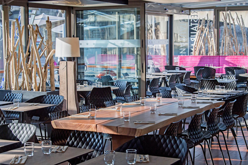 Modern restaurant with wooden tables, black quilted chairs, clear glasses, and silverware neatly arranged, next to large windows.