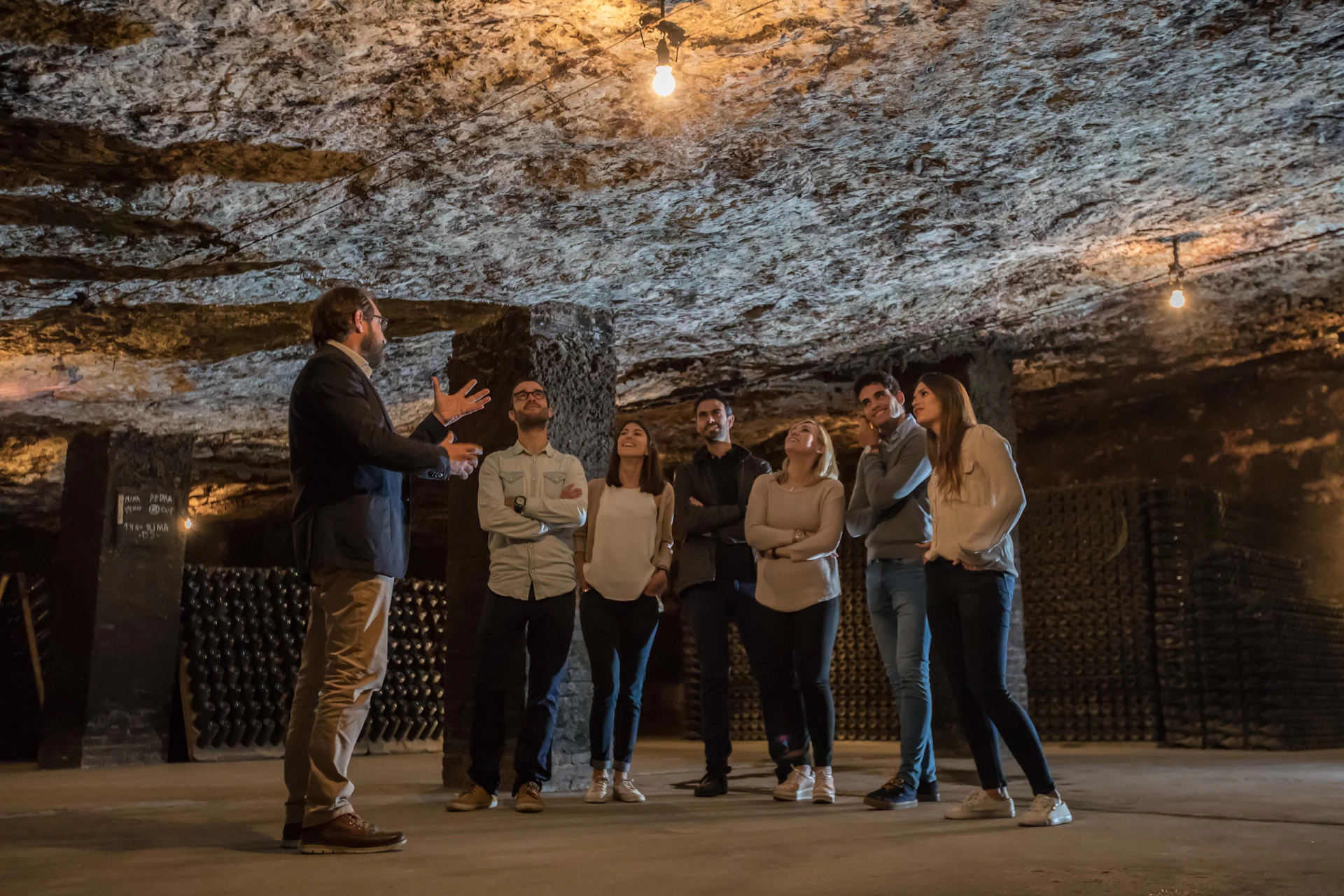 A group of seven people stand in a stone-walled room, attentively listening to a man speaking under a single hanging light bulb.