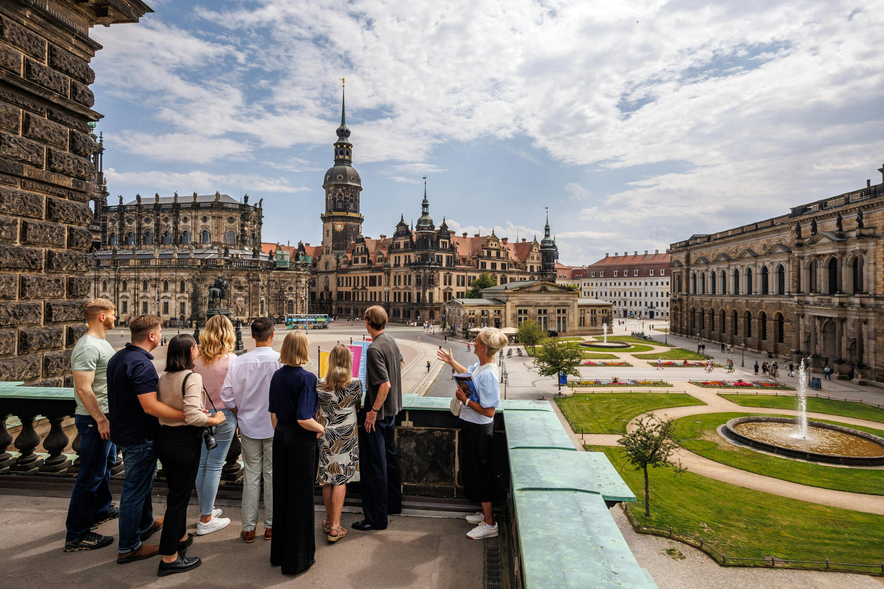 Un gruppo di persone è in piedi su una terrazza che si affaccia su una piazza storica del centro, circondata da edifici decorati, sotto un cielo parzialmente nuvoloso.