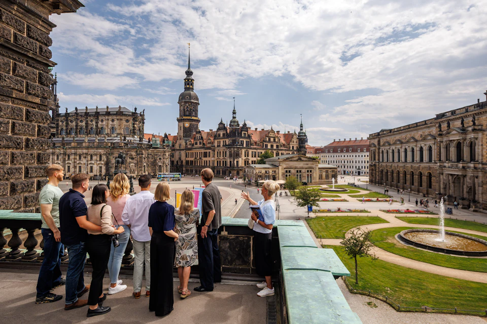 A group of people are standing on a terrace overlooking a historic town square with ornate buildings, beneath a partly cloudy sky.