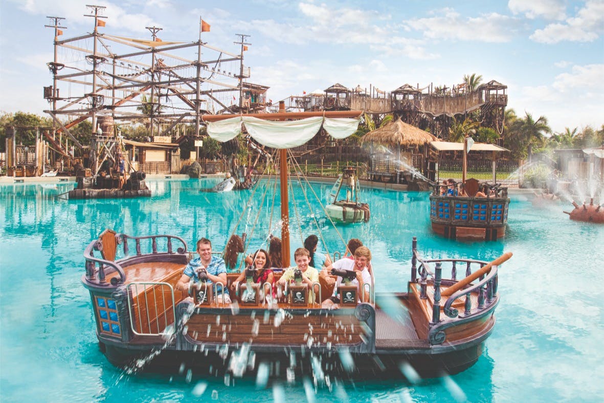 People enjoying a ride on a pirate-themed boat in a water park, with a ropes course structure visible in the background.
