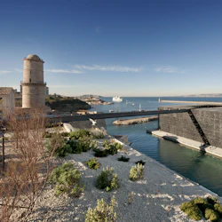 Havsutsikt med ett gammalt stentorn, en modern byggnad med invecklad design, gångbro, växter och ett skepp på vattnet.