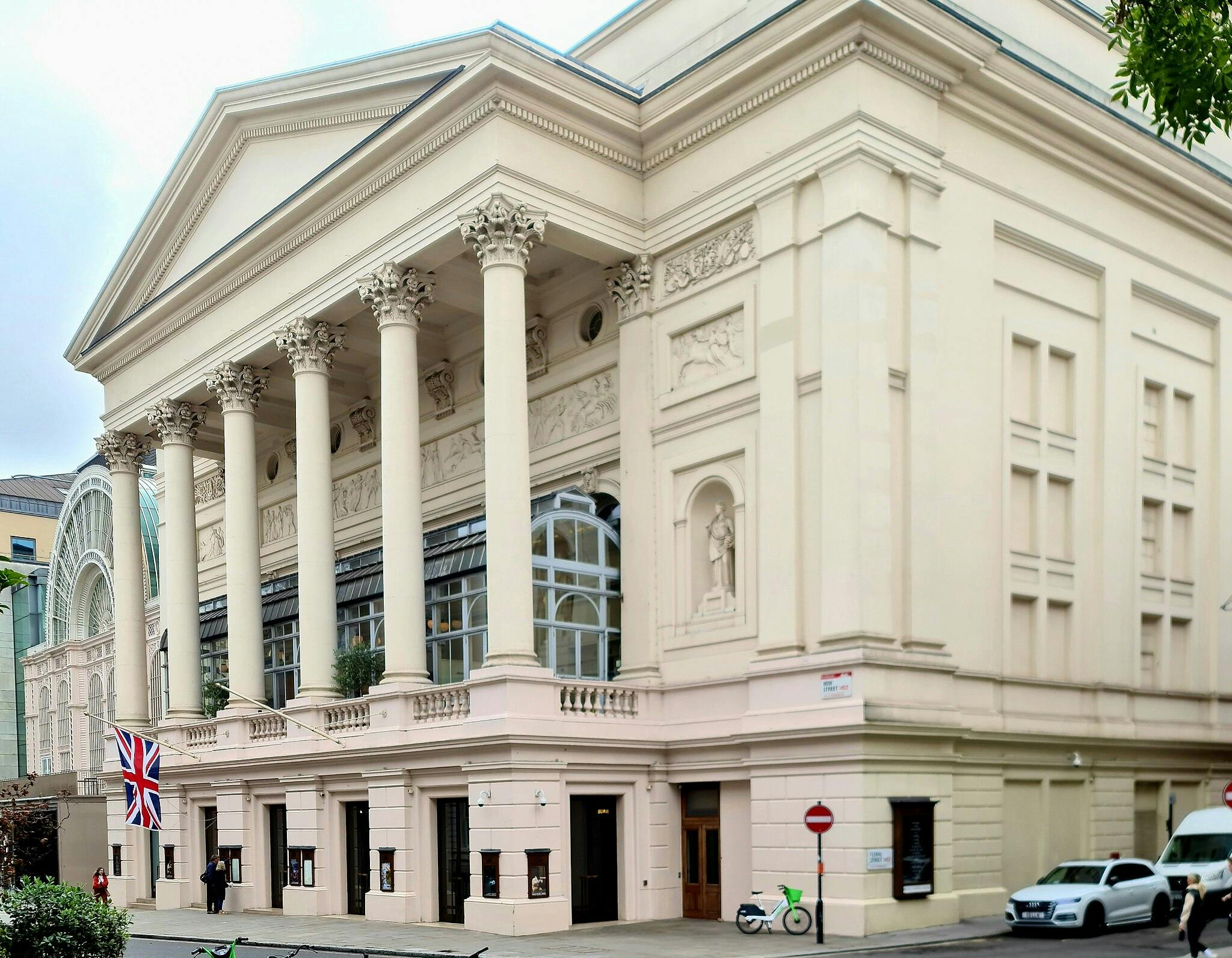 높은 코린토스 기둥, 디테일한 프리즈, 유니언 잭, 아치형 창문, 주차된 차량이 있는 신고전주의 양식의 건물입니다.